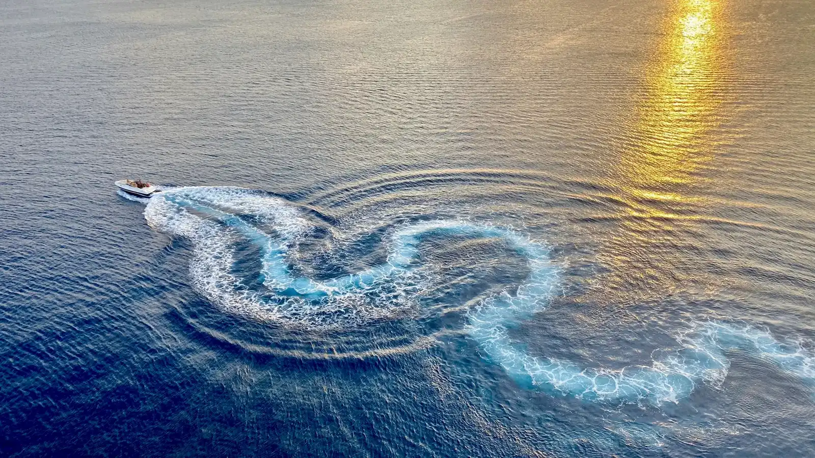 Aerial view of a speedboat carving a looping wake across open water as the late sun glints on the surface
