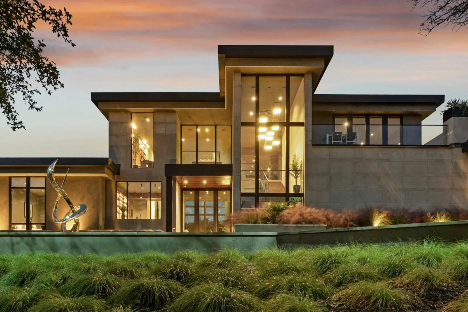 A modern luxury home with tall glowing windows at dusk above a planted lawn